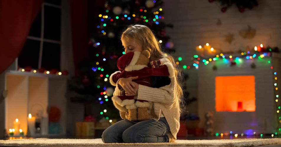 This picture shows a young girl hugging a teddy bear in front of a Christmas tree. Photo Courtesy: AMA Queensland 